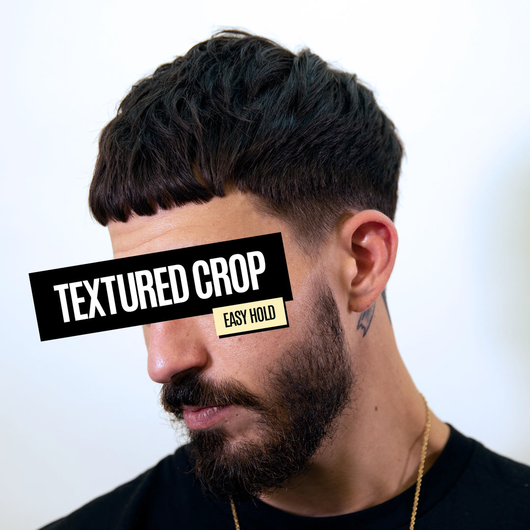 A close-up portrait of a man with short, textured dark hair, a beard, and a black crew cut, wearing a black shirt and gold chain. A black text overlay reads 'TEXTURED CROP' and a smaller yellow label says 'EASY HOLD' against a plain light background.